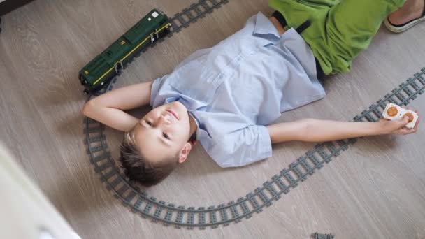 En tonåring leker med ett tåg på kontrollerna på golvet i rummet. Ovanifrån. Leksaksjärnväg. leende vänder han huvudet mot det rörliga loket — Stockvideo