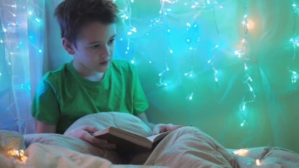 Ein Junge liest ein Buch, sitzt auf einem Bett, das mit Girlanden geschmückt ist. buntes Licht der Lichter. Ein Teenager sitzt unter einer Decke — Stockvideo