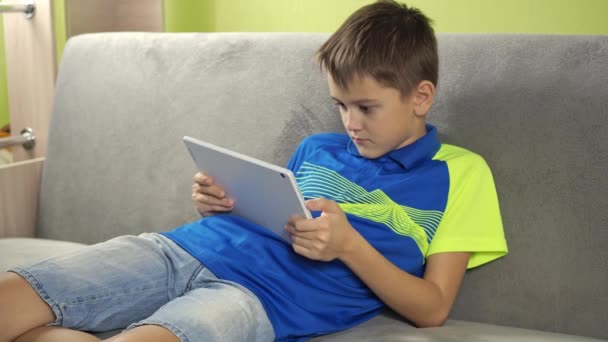 Подросток играет с планшетом. улыбка на его лице. мальчик в футболке и шортах сидит на диване в комнате — стоковое видео
