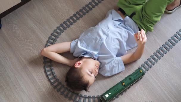 Ein Junge spielt mit einer Eisenbahn auf dem Boden im Raum. Ansicht von oben. Spielzeugeisenbahn. Lächelnd wendet er seinen Kopf der fahrenden Lokomotive zu — Stockvideo