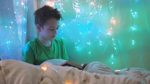 Um menino está lendo um livro sentado em uma cama decorada com guirlandas. luz multicolorida de luzes. um adolescente está sentado sob um cobertor — Vídeo de Stock