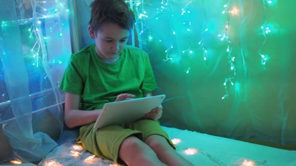 A boy is playing a tablet sitting on a bed decorated with garlands. multicolored light of lights. a teenager in a green T-shirt and shorts Video Clip