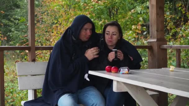 Молодая пара пьет чай, сидя на улице под одеялом и в теплой одежде. осень. греющие напитки — стоковое видео