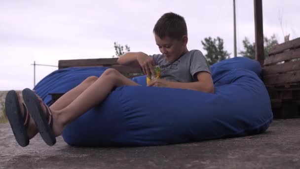 Un niño feliz come galletas acostado en una silla de bolsa. un sofá suave en la calle. el placer de la relajación — Vídeo de stock