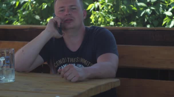 Egy férfi ül az árnyékban egy pavilonban egy asztalnál. Vedd fel a telefont. a gyerekes beszélgetésben. — Stock videók
