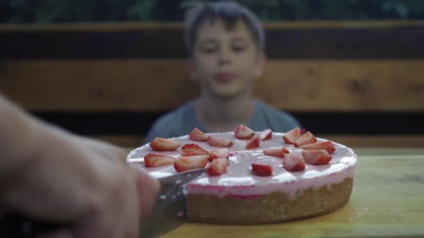 Eine Hand schneidet einen hausgemachten Erdbeerkuchen. Ein Junge beobachtet den Prozess. Der Junge ist aus dem Fokus, der Kuchen steht im Fokus — Stockvideo