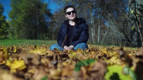 En ung kvinna sitter på en lövmatta. Kastar upp bladverket. poserar för en fotografering — Stockvideo