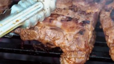 Domuz pirzolası ızgarada maşayla çevirilir. Kömürden çıkan duman. Izgarada yemek pişirmek. Yakın plan. Bir parça lezzetli baloncuğu çevirdikten sonra