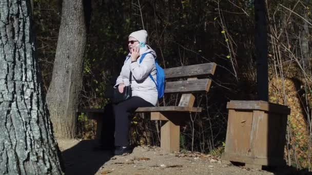 Uma mulher idosa está sentada em um banco de estacionamento e falando ao telefone. Outono no parque. ela está usando um casaco, um chapéu e óculos de sol. com um saco nas mãos — Vídeo de Stock