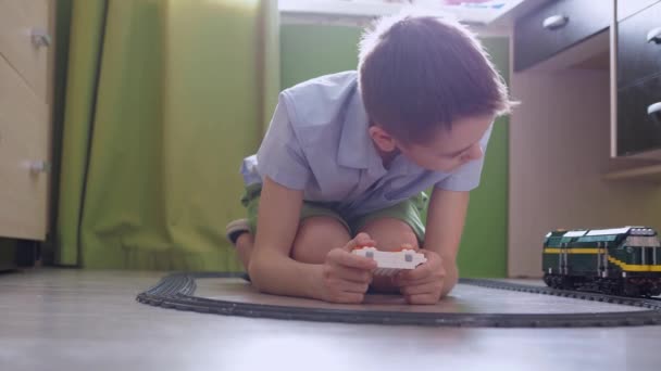 Un niño de 11 años está jugando con un tren en los controles en el suelo de la habitación. vista lateral — Vídeo de stock