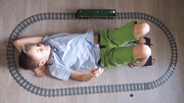 Un niño está jugando con un tren en los controles en el suelo en la habitación. Vista superior. ferrocarril de juguete. sonriendo, vuelve la cabeza hacia la locomotora en movimiento — Vídeos de Stock