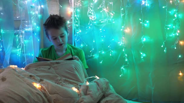 A boy is reading a book sitting on a bed decorated with garlands. multicolored light of lights. a teenager under a blanket Stock Footage