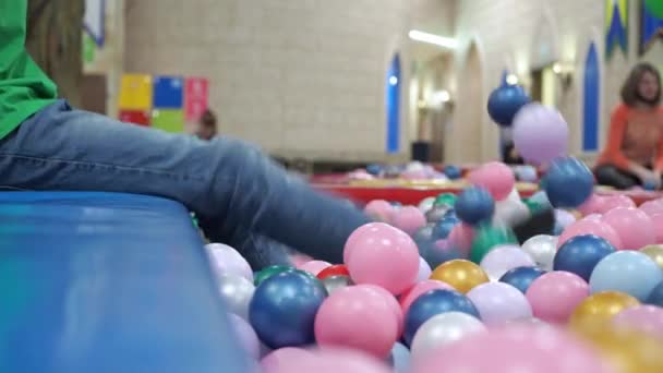 Un garçon joue avec ses pieds avec des balles dans une piscine avec des balles en plastique. centre de divertissement pour enfants — Video