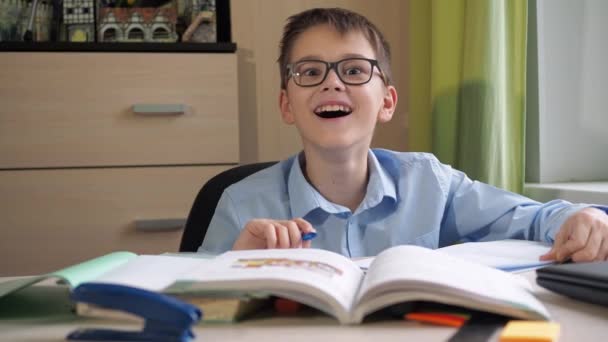 Ein Teenager mit Brille lächelt überrascht in die Kamera. Das Lehrbuch liegt auf dem Tisch. einen Schreibtisch. Hausunterricht. Fernstudie — Stockvideo