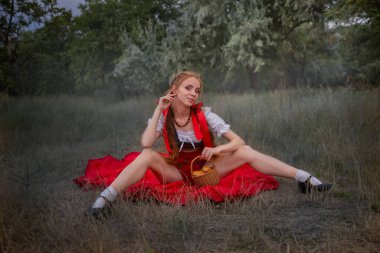 A cute fantasy image of a fairy tale character, a mysterious red-haired girl in the forest sits on a bright red bright cloak. Little Red Riding Hood in the forest - Portrait of the heroine of a children's book in the forest