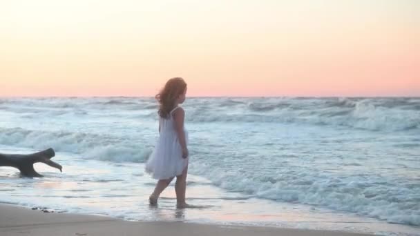 Holčička dítě v dlouhých slunečních šatech s dlouhými vlasy běží podél pláže podél moře při západu slunce v létě — Stock video