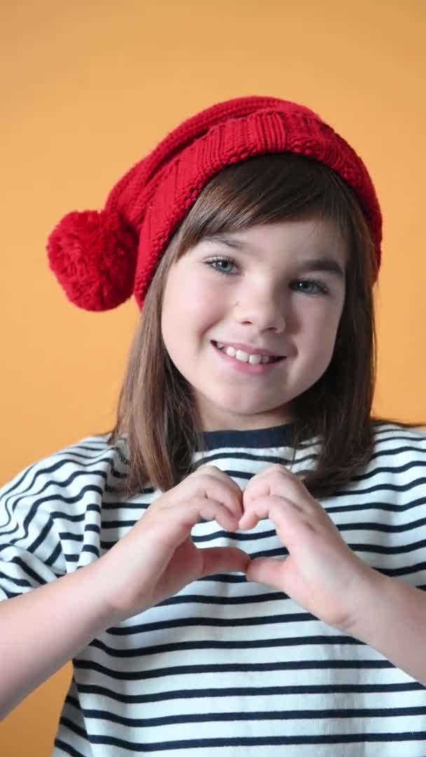 Χαμογελαστό κορίτσι στο καπέλο santa που δείχνει την καρδιά σε απομονωμένο κίτρινο φόντο — Αρχείο Βίντεο