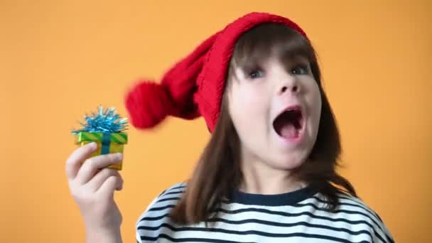 산타 모자를 쓴 귀여운 소녀가 노란 배경을 가까이 서 보고 는 감정적으로 기뻐 한다 — 비디오