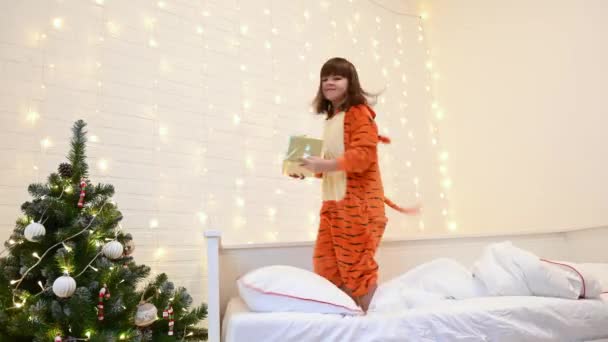 Ένα κορίτσι ντυμένο τίγρη ως σύμβολο του 2022 κοιτάζει ένα δώρο στο κρεβάτι δίπλα σε ένα χριστουγεννιάτικο δέντρο — Αρχείο Βίντεο