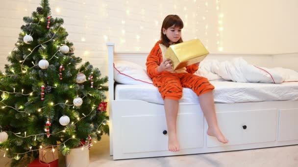 2022 'nin sembolü olarak kaplan gibi giyinmiş bir kız Noel ağacının yanındaki yatakta hediyeye bakar. — Stok video