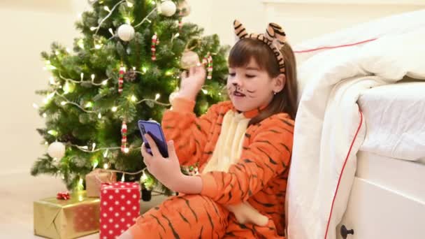 Ένα κορίτσι σε ένα κοστούμι τίγρης δίπλα σε ένα χριστουγεννιάτικο δέντρο περιμένει τα Χριστούγεννα και τα δώρα που επικοινωνούν σε ένα smartphone μέσω βίντεο επικοινωνίας — Αρχείο Βίντεο
