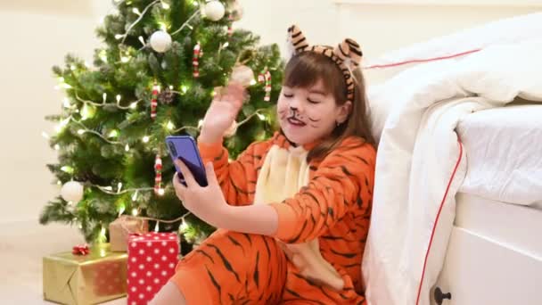 クリスマスツリーの横にあるトラの衣装を着た女の子がクリスマスを待っていて、ビデオ通信でスマートフォンで通信する贈り物を待っています — ストック動画