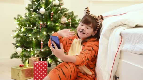 Девушка в костюме тигра рядом с рождественской ёлкой ждет Рождества и подарков, общающихся на смартфоне через видеосвязь — стоковое видео