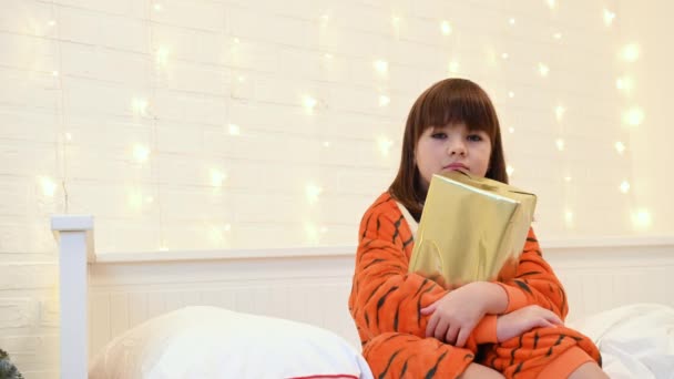 Uma menina vestida de tigre como um símbolo de 2022 olha para um presente na cama ao lado de uma árvore de Natal — Vídeo de Stock