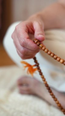 dikey video. Bir adam lotus pozisyonunda oturur. Tespihi parmaklayan ellerin yakın çekimi. izoterik tema