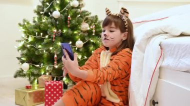 Noel ağacının yanında kaplan kostümlü bir kız Noel 'i bekliyor ve video iletişimiyle akıllı telefondan iletişim kuruyor.