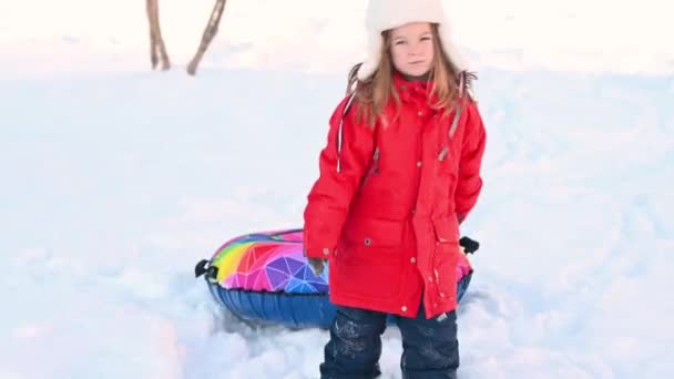 Niña divertida en una chaqueta roja con un tubo inflable cabalga por un tobogán en la nieve en invierno — Vídeo de stock