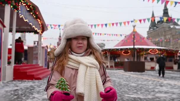Россия Москва Люди ходят по красной площади, маленькая смешная девочка ест пряник на палочке на фоне Рождественской ярмарки и карусели зимой — стоковое видео