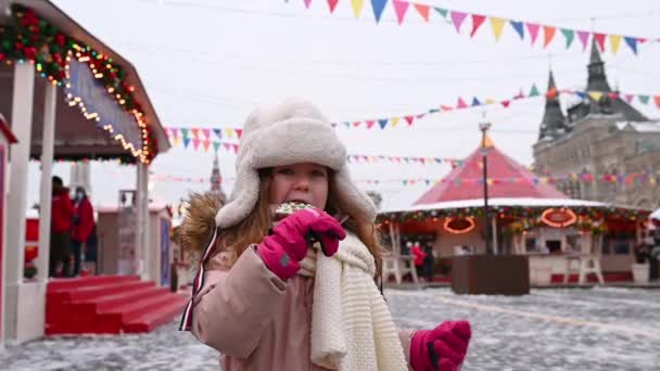 Московські люди ходять на червоній площі, маленька кумедна дівчинка їсть пряник на паличці на фоні Різдвяного ярмарку і карусель взимку. — стокове відео