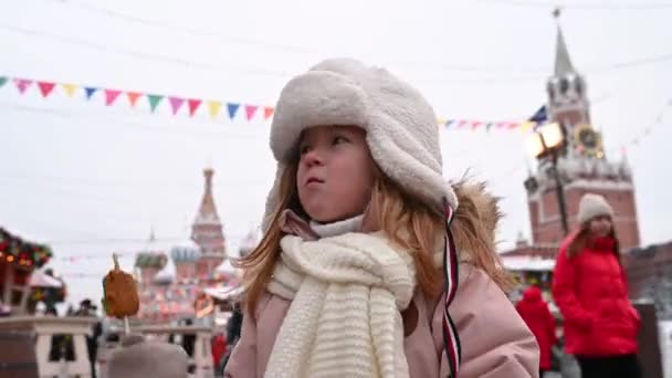 Russie Moscou Les gens marchent sur la place rouge, une petite fille drôle mange un pain d'épice sur un bâton sur le fond d'une foire de Noël et un carrousel en hiver — Video