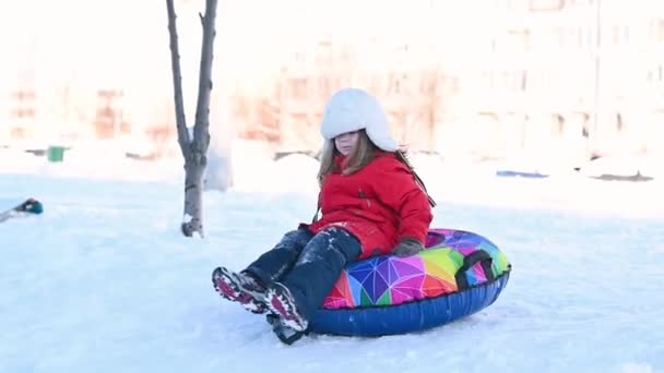 Маленькая забавная девочка в красной куртке с надувной трубкой едет вниз по горке в снегу зимой — стоковое видео