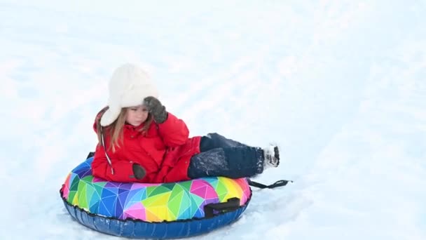 Маленькая забавная девочка в красной куртке с надувной трубкой едет вниз по горке в снегу зимой — стоковое видео