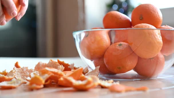 Ljusa orange tangeriner ligger på bordet, flickan tar en mandarin och skalar huden mot bakgrund av blinkande julbelysning — Stockvideo