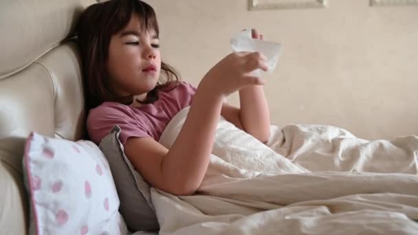 A 10-year-old girl lies in bed sick, coughs, blows her nose into a napkin — Stock Video