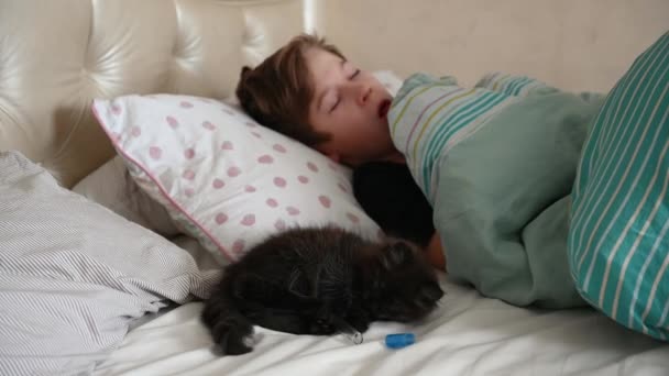 12-letý chlapec leží v posteli nemocný, kašle, vypadá to, že si hraje s teploměrem na posteli legrační černé kotě — Stock video