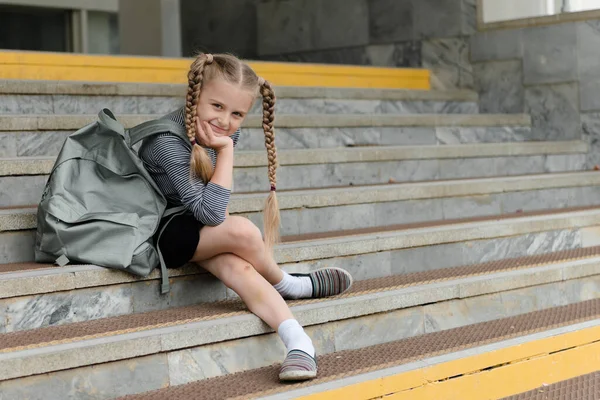 Милая счастливая девочка школьница с рюкзаком сидя рядом со школьной темой образования — стоковое фото