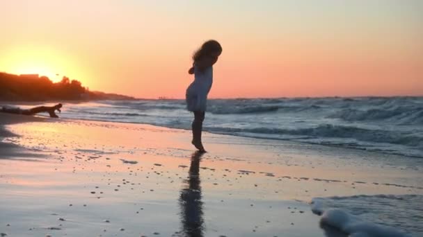 長い髪をした長いサンドレスを着た少女が夏の日没に海沿いのビーチを走り — ストック動画