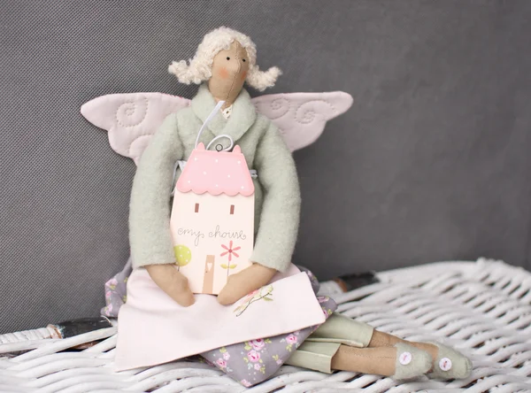 Tilda boneca anjo whith pequena casa em braços sentado no peito de vime em fundo cinza — Fotografia de Stock