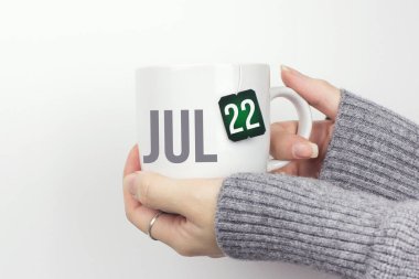 22 Temmuz. Ayın 22 'si, takvim tarihi. Gri kazaklı kadınların elinde çay fincanı ve çay poşetindeki takvim tarihli çay fincanı var. Yaz ayı, yılın günü konsepti.