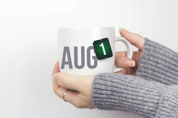 8月1日 月の1日 カレンダーの日付 グレーのセーターに身を包んだ女性の手を閉じ ティーカップを持ち ティーバッグのラベルに月とカレンダーの日付を記入します 夏の月 その年のコンセプトの日 — ストック写真