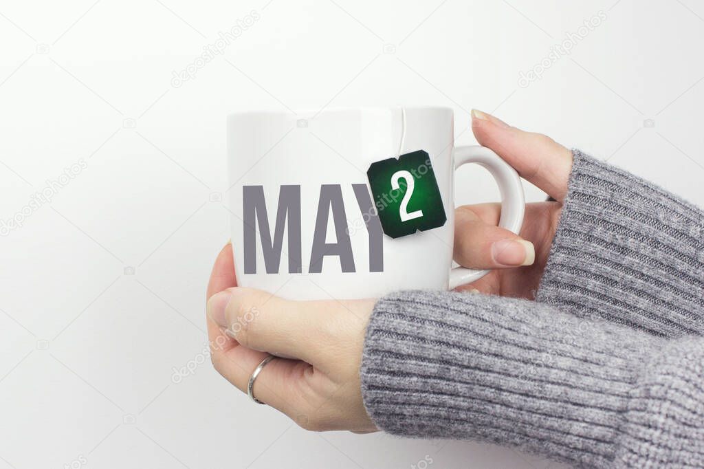 May 2nd. Day 2 of month, Calendar date. Closeup of female hands in grey sweater holding cup of tea with month and calendar date on teabag label. Spring month, day of the year concept