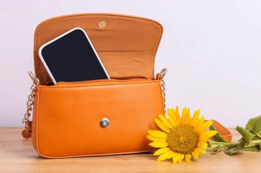 Orange women's crossbody bag with smartphone inside and sunflower on wooden background.