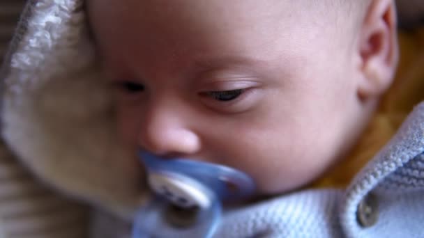 Look of baby in camera close up shot. infant, childhood, parental love, emotion concept - cute smiling face of brown-eyed chubby newborn with pacifier dummy in soft blue jacket awake looks around home — Stock Video