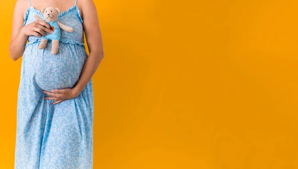 Macierzyństwo, kobiecość, witaminy, gorące lato - portret ciężarnej kobiety w kwiatowej niebieskiej sukience posiada miękki pluszowy miś głaskający brzuch dłonią na żółtym tle przestrzeń do kopiowania — Zdjęcie stockowe