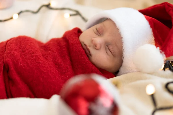 Top View Portrait First Days Of Life Newborn Cute Funny Sleeping Child Baby In Santa Hat Wrapped In Red Diaper At White Garland Background. Merry Christmas, Happy New Year, Infant, Childhood, Winter