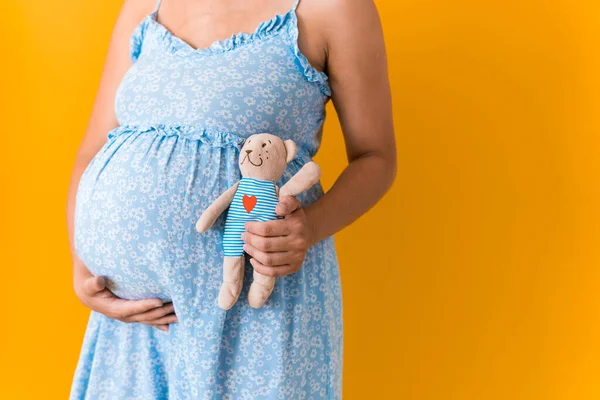 Maternidade, feminilidade, verão quente - retrato cortado grávida mulher irreconhecível em vestido azul contém brinquedo macio ursinho acariciando barriga esperando bebê no amarelo cor brilhante fundo espaço de cópia — Fotografia de Stock